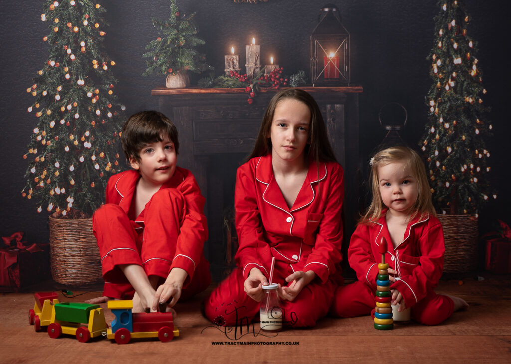 Three siblings in matching Christmas red pyjamas sitting on the floor in front of fire