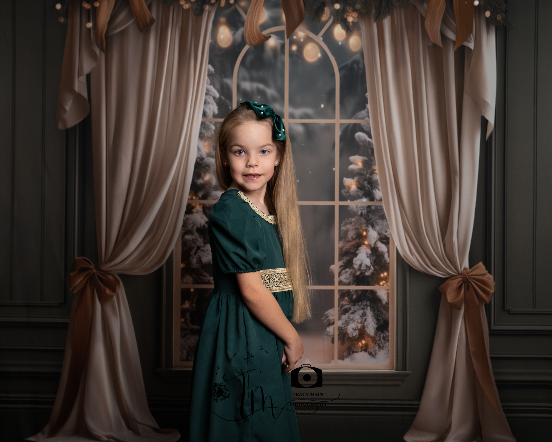 TMP_5213-Edit Girl with long blonde hair in a green party dress in front of a window with snow outside and Christmas lights on the trees